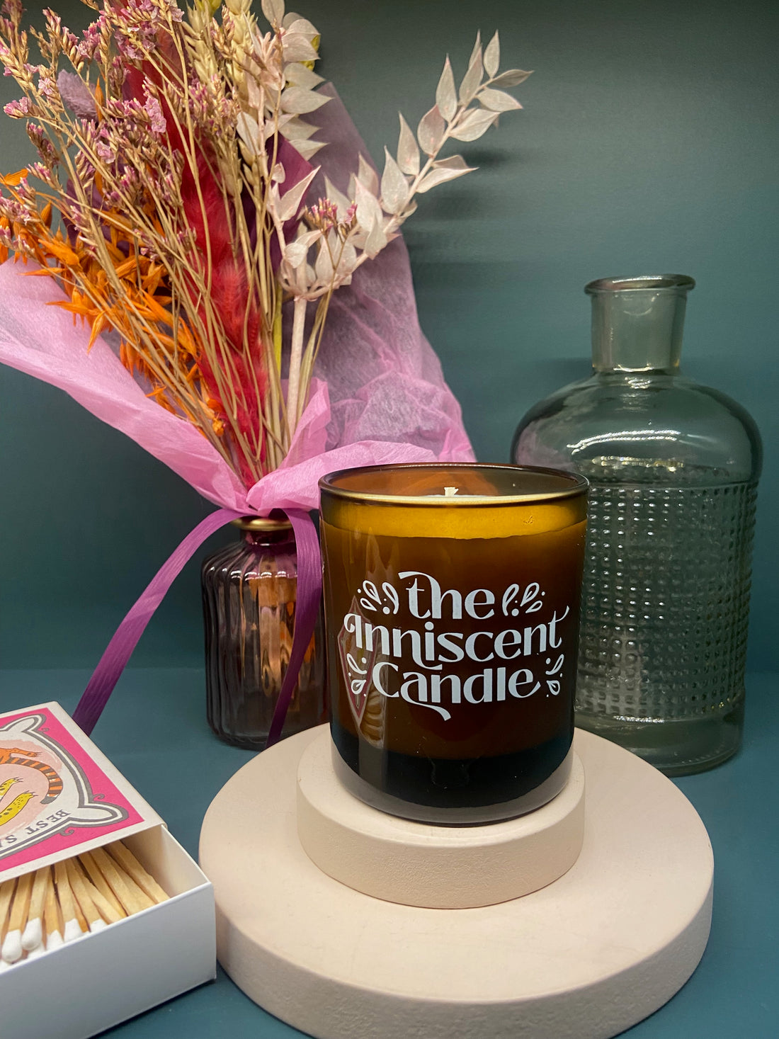 Image of a hand poured candle, sitting on a pink pedestal, with a bunch of colourful flowers in the background. Glass bottle and box of matchsticks are also present.
