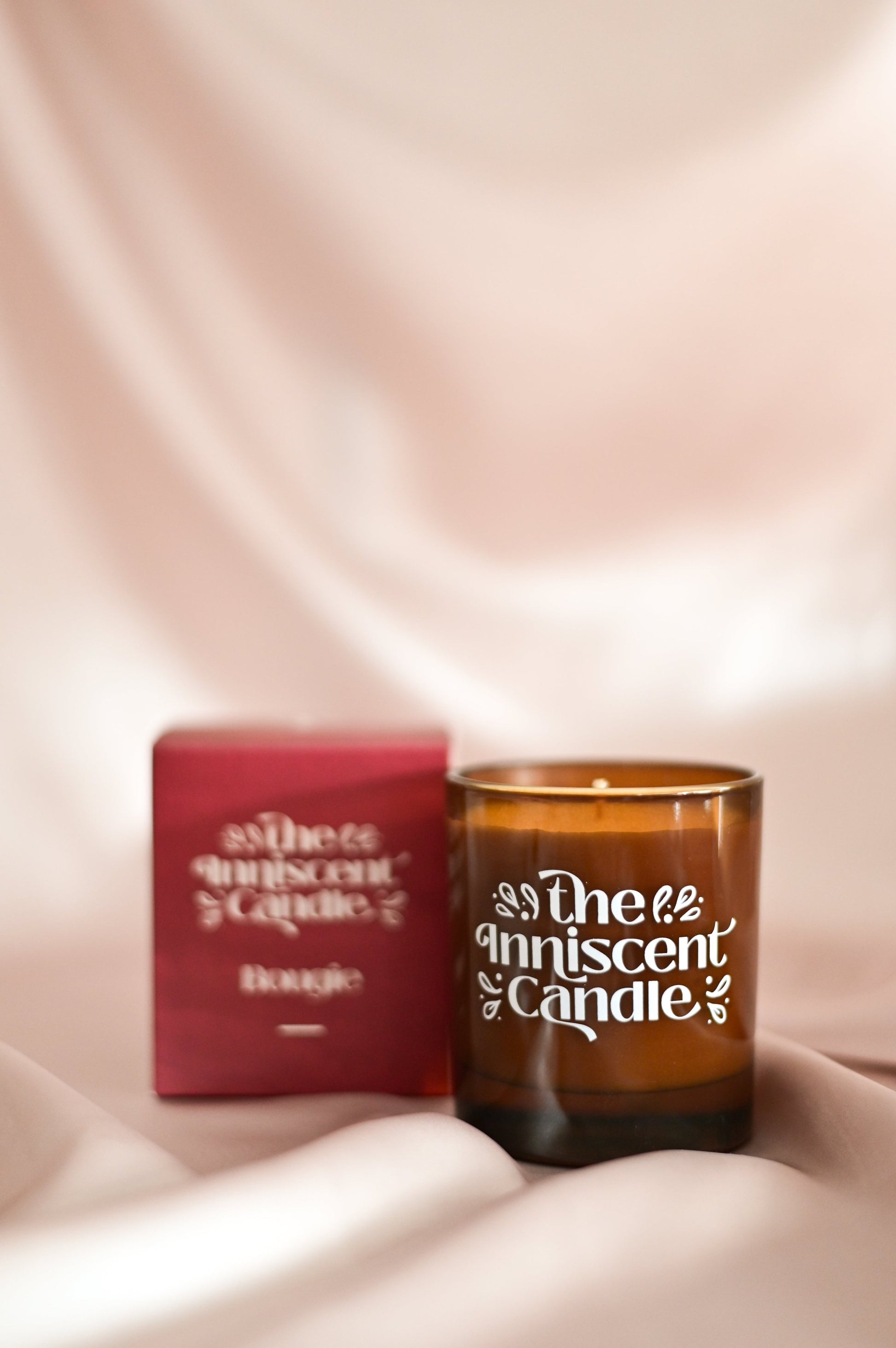 Photo of a soy candle in amber glass, beside box, on a silky champagne coloured fabric.