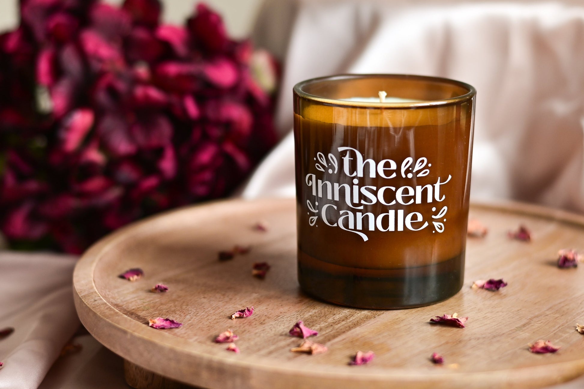 Photo of a soy candle in amber glass, with dried deep pink florals, on an oak stand.