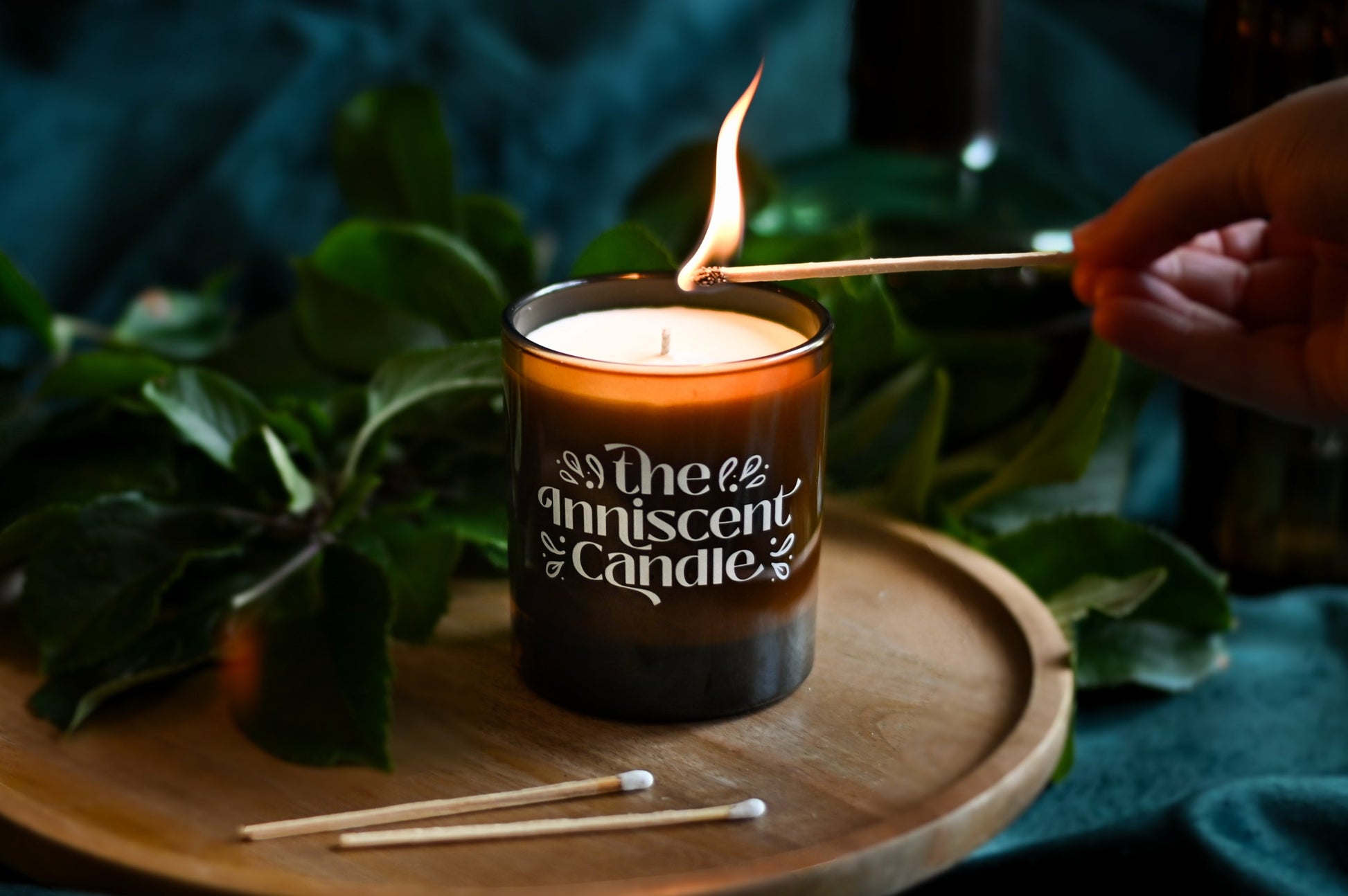 Photo of a soy candle in amber glass, being lit with a match with greenery in background.