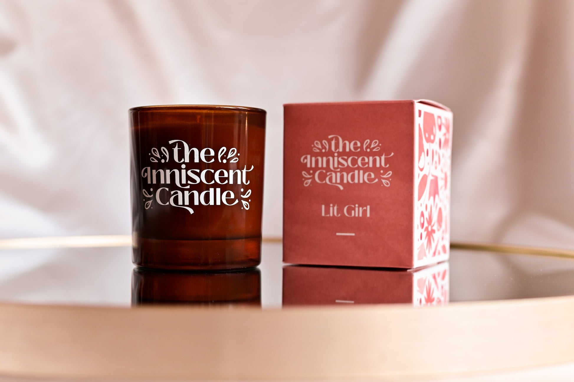 Photo of a soy candle in amber glass, with box on a mirrored surface, set against silky champagne-coloured fabric.