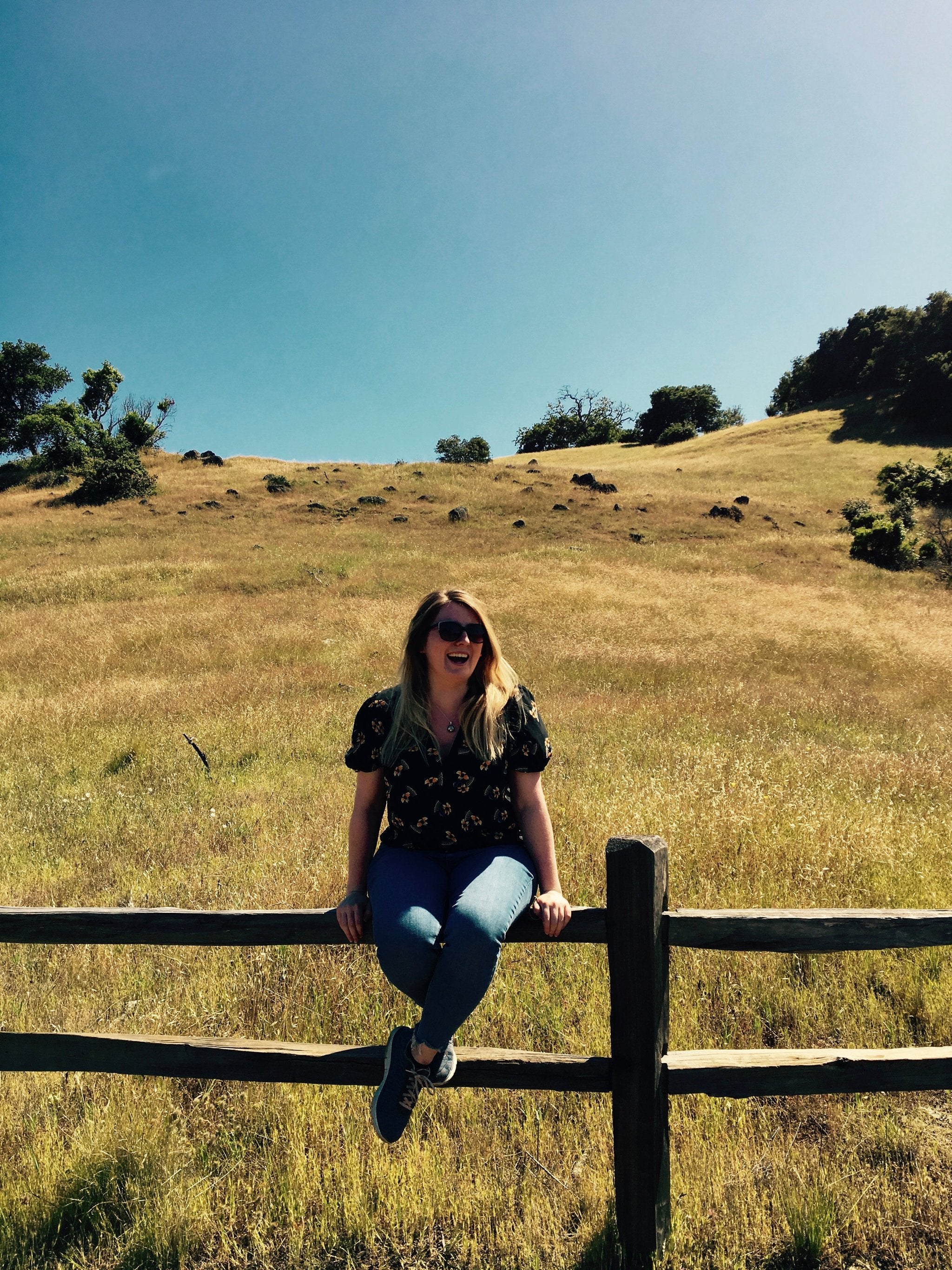 The Inniscent Candle Founder, Rebecca, perched on a fence with rolling hills in the background.